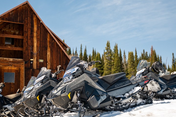 Snowmobiles parked in snow near a rustic wooden cabin with a forest in the background.