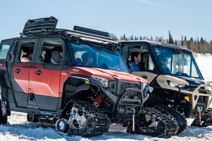 Two tracked off-road vehicles driving on snow in a forested area.