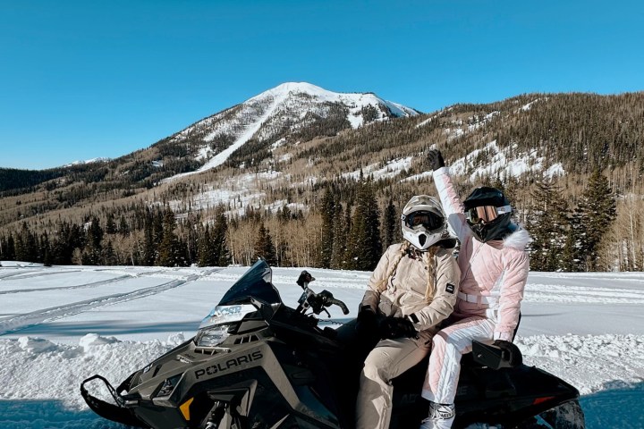 Two people in winter gear on a snowmobile with a snowy mountain backdrop.
